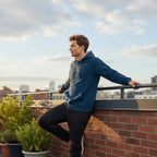 Man in a blue hoodie leaning against a railing on a rooftop with a cityscape in the background