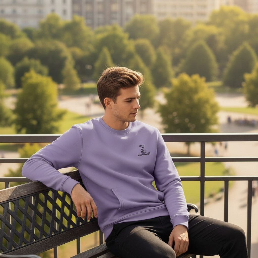 Man wearing a purple sweatshirt with a logo, sitting on a bench in an outdoor setting with trees and buildings in the background.