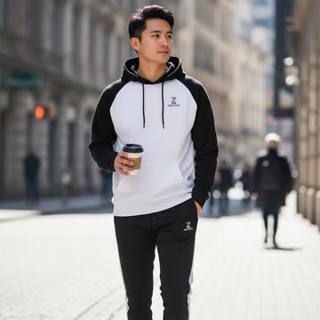 Man walking on a city street wearing a black and white hoodie with a logo, holding a coffee cup.