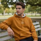 Man wearing a mustard yellow hoodie with a logo, sitting on a bench in a park.