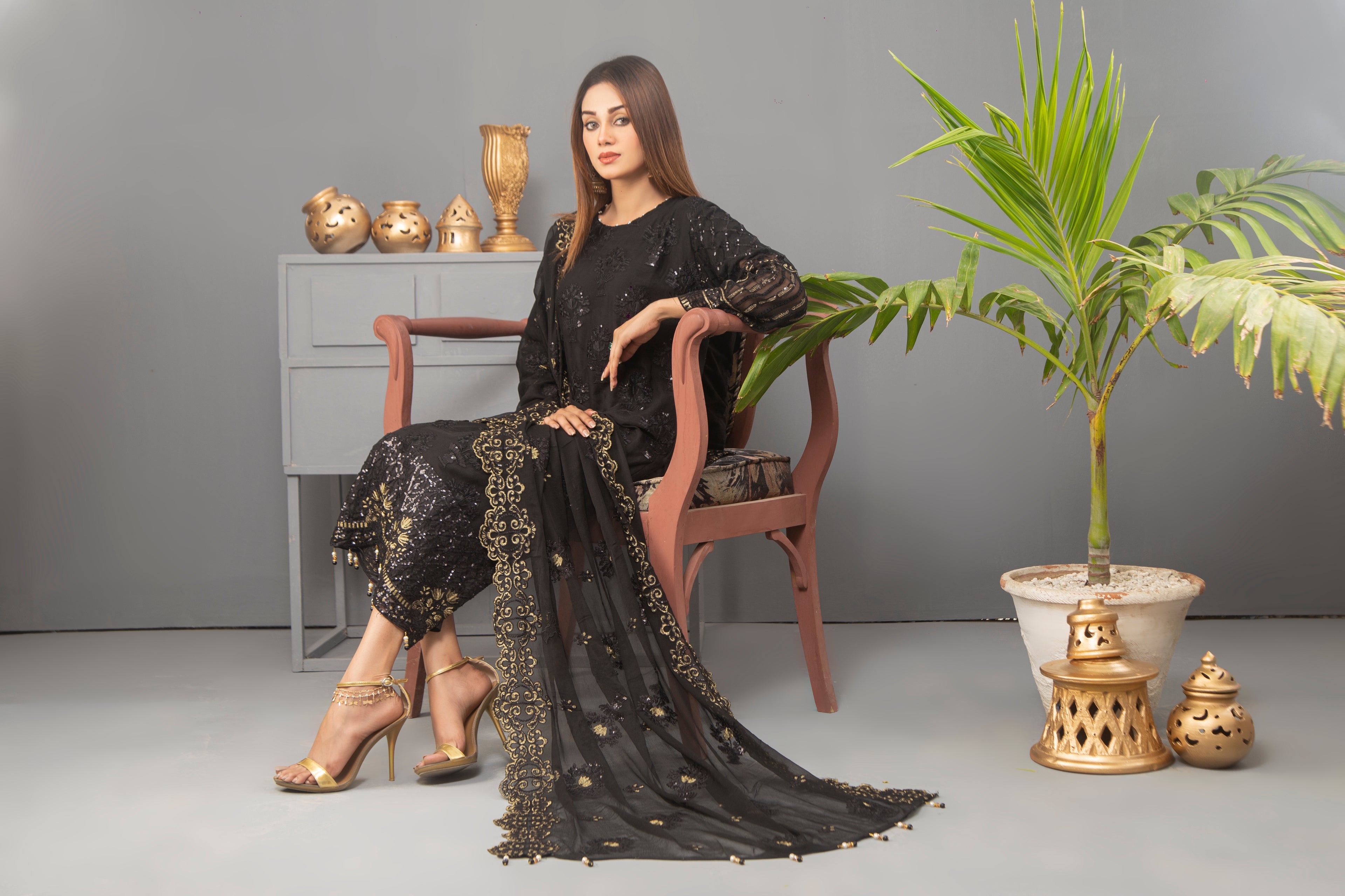 Woman in a black embroidered outfit sitting on a chair with decorative items and plants in the background.